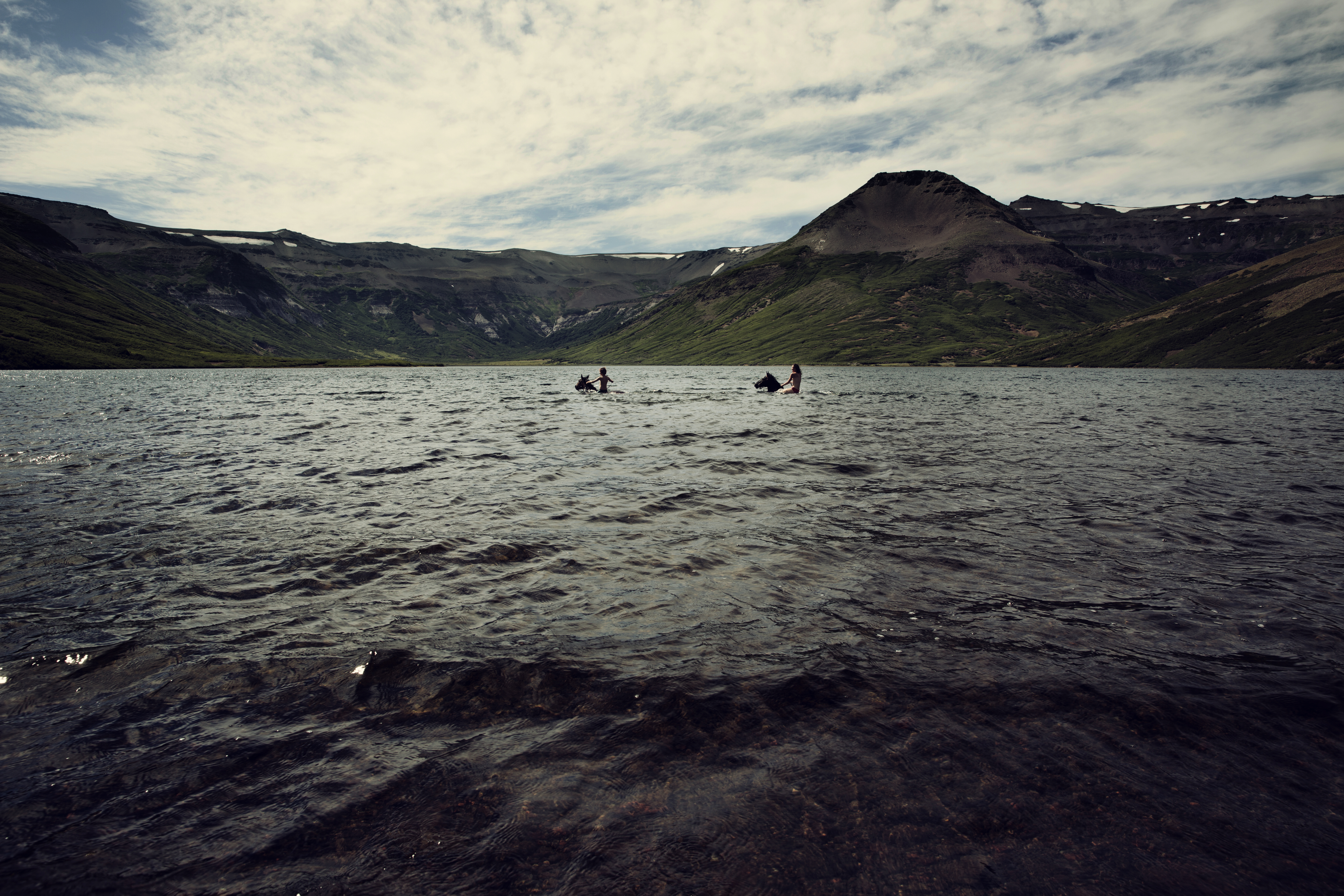 Two people swimming on horses in alpine lake on horse vacation at Estancia Ranquilco in Patagonia Argentina 